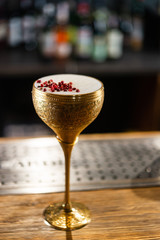Cocktail with white foam decorated with red dried peppers served in the metal gold glass on the wooden bar table
