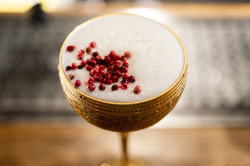 Cocktail with white foam decorated with red dried peppers served in the metal gold glass on the wooden bar table