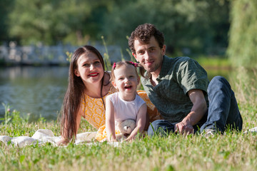 Naklejka na ściany i meble Happy family relaxing in the Park. They are lying on a white plaid. Girl holding in hand a Teddy bear. In the background the lake.