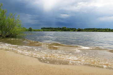 The sandy shore of the lake is covered with large waves and a pre-storm sky.