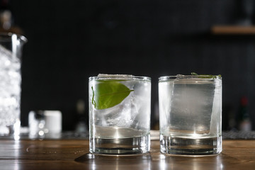 Transparent cocktail in rocks glass decorated with one big ice cube and green leaf served on the wooden bar desk.
