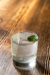 Transparent cocktail in rocks glass decorated with one big ice cube and green leaf served on the wooden bar desk.