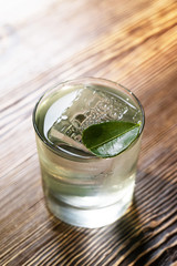 Transparent cocktail in rocks glass decorated with one big ice cube and green leaf served on the wooden bar desk.