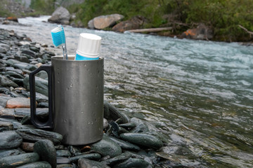 Camping mug with toothbrush and toothpaste on the bank of a mountain river. Do not forget about hygiene while hiking in the mountains