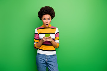 Photo of pretty blogger dark skin curly lady holding telephone hands reading awful fake news about herself open mouth wear casual striped pullover jeans isolated green color background