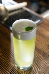 Green cocktail on bar counter. Drink with martini, gin, rum, tequila with alcohol and white foam on the top decorated with a basil leaf. Glass with booze on a wooden board.