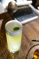 Green cocktail on bar counter. Drink with martini, gin, rum, tequila with alcohol and white foam on the top decorated with a basil leaf. Glass with booze on a wooden board.