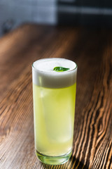 Green cocktail on bar counter. Drink with martini, gin, rum, tequila with alcohol and white foam on the top decorated with a basil leaf. Glass with booze on a wooden board.