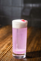 Space violet alcoholic drink in the big cocktail glass decorated with a red flower on the wooden bar desk counter under the studio lights