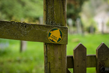 Sign: Public Footpath, seen on a wooden fence, with a blurry cemetery in the background, seen in Aysgarth, North Yorkshire, England, UK