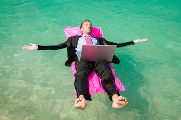 Barefoot businessman floating outdoors with laptop computer on a pink inflatable lilo in bright...