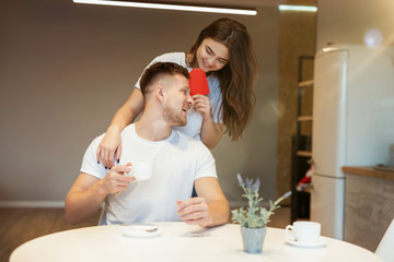 Obraz na płótnie Canvas young beautiful woman surprising her husband with paper heart on saint valentine's day during morning coffee in the kitchen, happy romantic unniversary