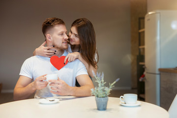 Obraz na płótnie Canvas young beautiful woman hugging and surprising her husband with red paper heart on saint valentine's day during breakfast in the kitchen, happy romantic unniversary