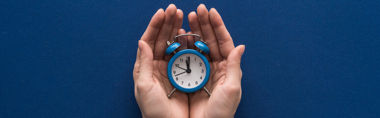 cropped view of woman holding alarm clock on blue background, panoramic shot