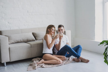 Young couple sit on the floor in stylish loft minimalist interior near the sofa and large window on the plaid and toasting champagne