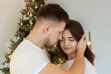 lovely couple husband hugging his beautiful wife standing near christmas tree both happy in room decorated for celebrating new year festive mood love story