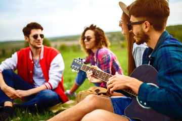 Group of happy friends with guitar having fun spending free time together in park sitting on grass. The guy plays the guitar. Young people enjoying party in the summer park. Rest, fun, summer concept.