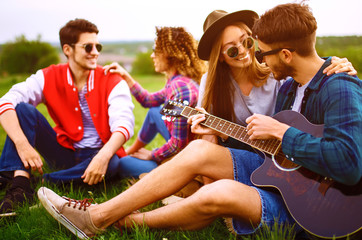 Group of happy friends with guitar having fun spending free time together in park sitting on grass. The guy plays the guitar. Young people enjoying party in the summer park. Rest, fun, summer concept.
