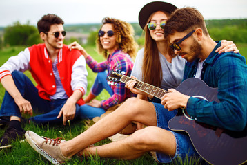 Group of happy friends with guitar having fun spending free time together in park sitting on grass. The guy plays the guitar. Young people enjoying party in the summer park. Rest, fun, summer concept.