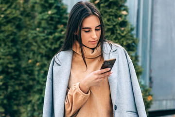 Attractive young woman using mobile phone and browsing online outdoor. Pretty brunette female standing in the city street calling her friend.