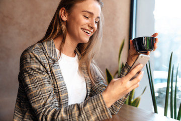 Photo of smiling young woman drinking coffee and using cellphone