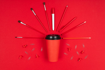 Brushes, paper clips, marker around a cup of coffee isolated on red background
