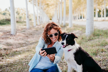 woman and beautiful border collie dog sitting in a path of trees outdoors. woman taking a selfie with mobile phone