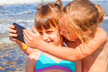 Summer holiday and leisure concept. Two cute child girls taking selfie together on smart phone on beach of sea.