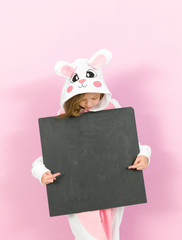 pretty blonde girl with cozy rabbit costume and blackboard is posing in the studio and is happy in front of pink background