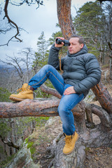 Traveler with monocle sitting on a large branch of conifer and looks into the distance.