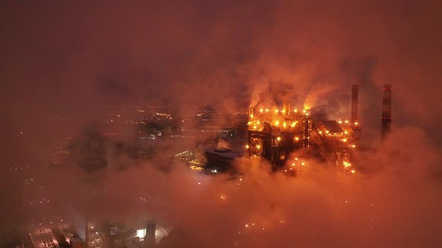 Aerial view of steel plant at night with smokestacks and fire blazing out of the pipe. Industrial panoramic landmark with blast furnance of metallurgical production. Concept of environmental pollution