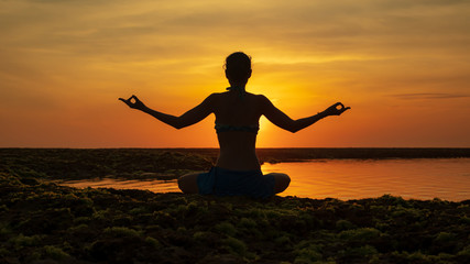 Yoga pose. Woman sitting on the beach, practicing yoga. Young woman raising arms with gyan mudra during sunset golden hour. View from back. Melasti beach, Bali.