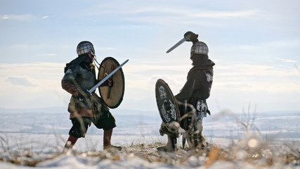 Two warriors viking are fighting with axes and shields on the winter meadow.