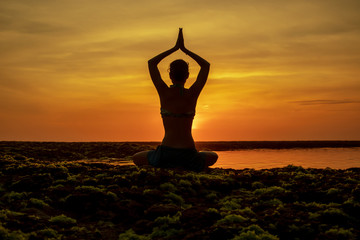 Yoga pose. Woman sitting on the beach, practicing yoga. Young woman raising arms with namaste mudra during sunset golden hour. View from back. Melasti beach, Bali. - obrazy, fototapety, plakaty