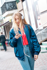 Outdoors Recreation. Teen girl student in denim jacket and headphones walking on urban background listening music browsing smartphone thoughtful