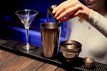 Girl bartender is preparing a cocktail by pouring an ingredient into an iron shaker.