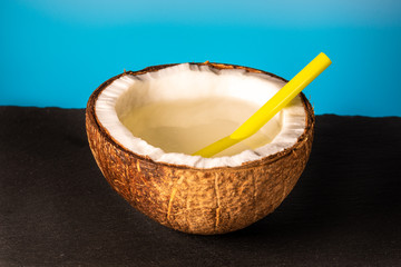 half of coconut with a plastic cocktail straw lies on a stone tray on a blue background