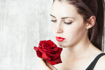 Beautiful young brunette Woman with red lips and red rose in her hand near the face as a symbol of love and care on St. Valentine's Day on the light gray concrete background with copy space