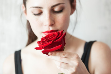 Beautiful young brunette Woman with red lips and red rose in her hand near the face as a symbol of love and care on St. Valentine's Day on the light gray concrete background with copy space