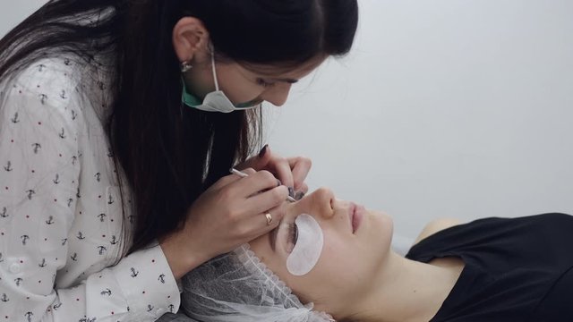 A brunette European lash artist in a disposable mask is doing lamination of the eyelashes. The client woman is lying on a couch during the procedure.The lash maker is brushing the lashes by a lash