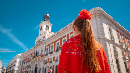 A young caucasian woman chulapa with red flower, traditional dress, and Spanish scarf at Puerta del Sol during San Isidro, the spring festival in May in the downtown of Madrid, the capital of Spain
