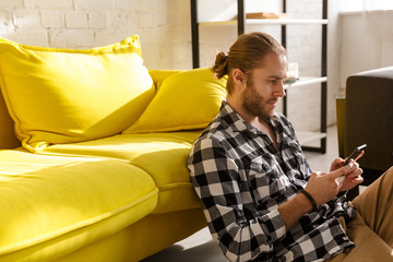 Photo of young man holding cellphone and sitting on floor in apartment