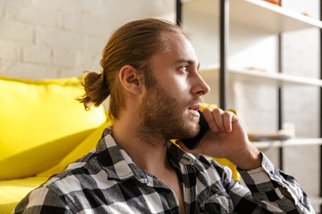 Photo of young man wearing plaid shirt talking on cellphone in apartment