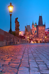 Charles Bridge at night with silhouette of a Saint, street light, historic buildings of Mala Strana, Lesser Bridge Tower and towers of Church of St. Nicolas Church and bellfry at dawn