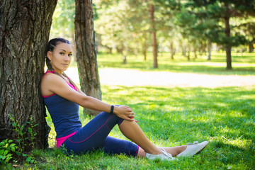 Sporty woman sitting on the grass leaning against a tree in the park on the background of trees