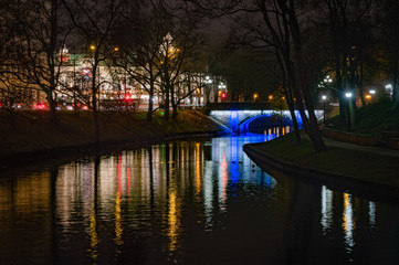 Fototapeta premium Riga's City Canal by night with the city lights reflecting in the water in foggy November evening and small bridge lightened with blue light and National Theater in background