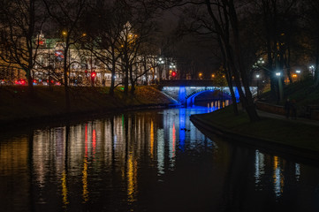 Small bridge lightened with blue light over Riga's City Canal by night with the city lights reflecting in the water in foggy November evening. National Theater in background