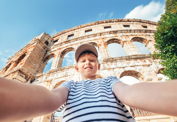 Young smiling teenager boy using two hands carrying a photo camera for taking a selfie portrait on the Pula city Amphitheatre ruins in Croatia. Famous landmarks traveling and vacation concept image.