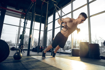 Overcoming. Young muscular caucasian athlete training in gym, doing strength exercises, practicing, work on his upper body with weights rings. Fitness, wellness, sport, healthy lifestyle concept.