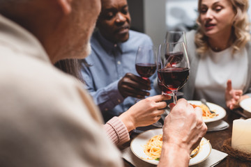 cropped view of multicultural friends talking and holding wine glasses during dinner